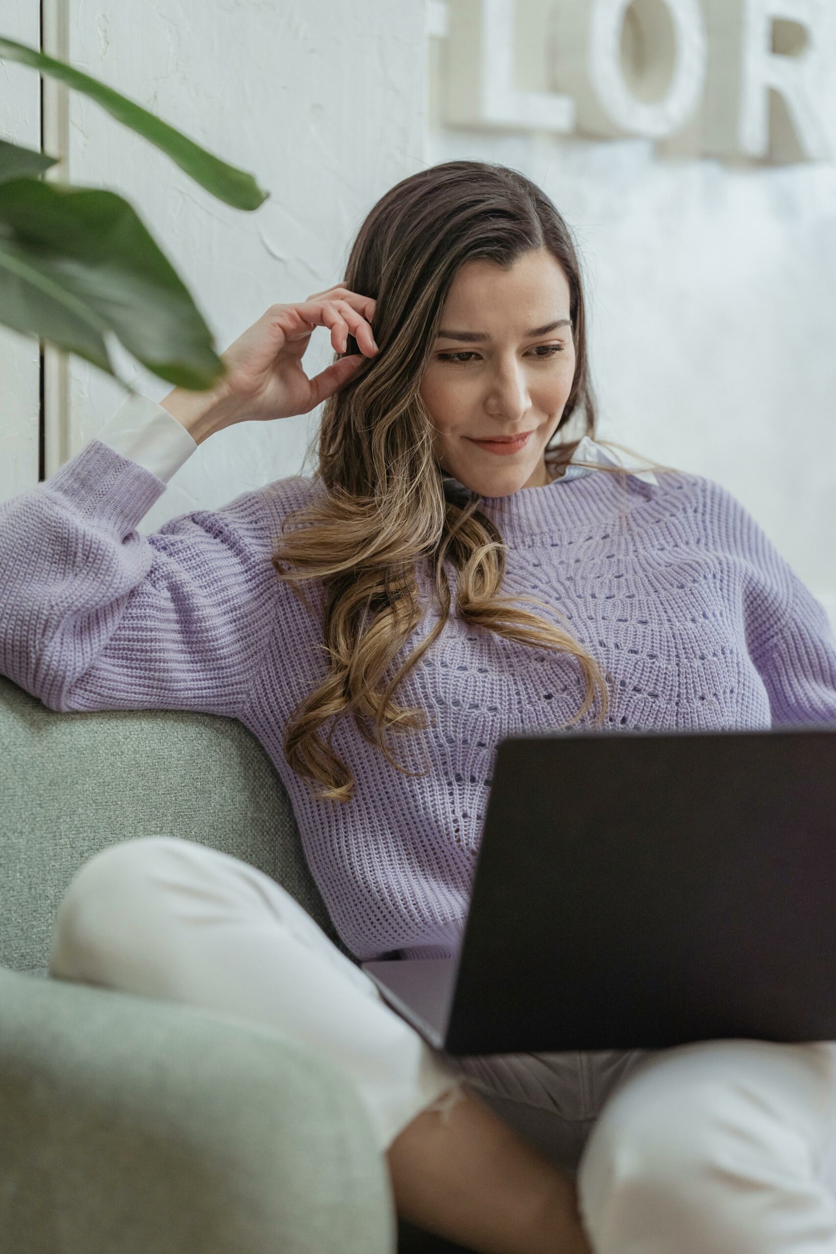 Young content female wearing casual clothes browsing netbook and touching hair while sitting on cozy sofa